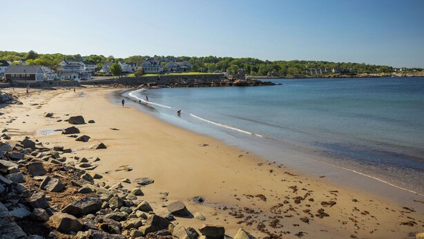 Une plage au rivage rocheux, avec une eau claire et des maisons en arrière-plan.