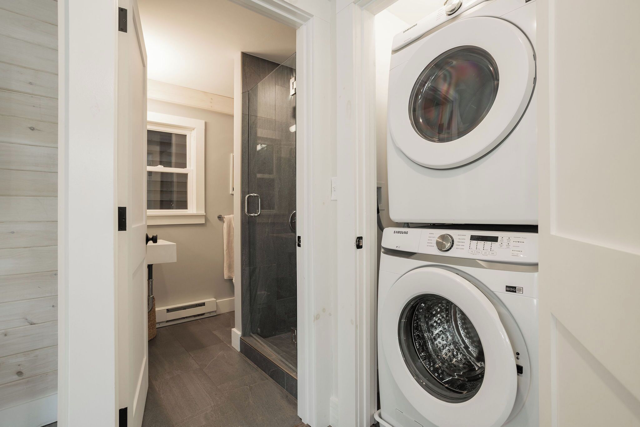 A laundry room with two stacked washing machines, a glass shower enclosure, and a small window.