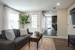 A living room with a sofa, a coffee table, a plant, and a view into the kitchen.