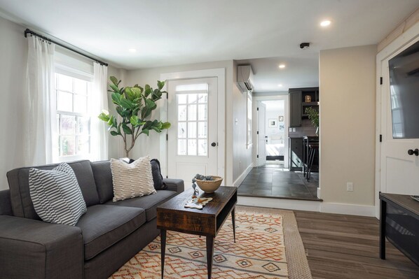 A living room with a sofa, a coffee table, a plant, and a view into the kitchen.