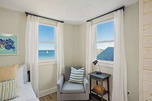 A corner bedroom with a view of the sea, a chair with a striped pillow, a bedside table with a lamp, and a painting on the wall.