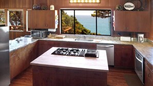 A modern kitchen with wooden cabinets, a central island with a stove, a large window with a view of the ocean, and a microwave on the countertop.