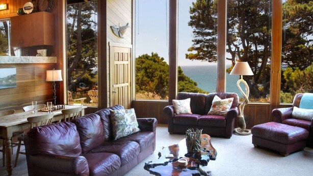 A spacious living room with a view of the ocean, featuring leather sofas, a coffee table, and a dining area with a chess set.