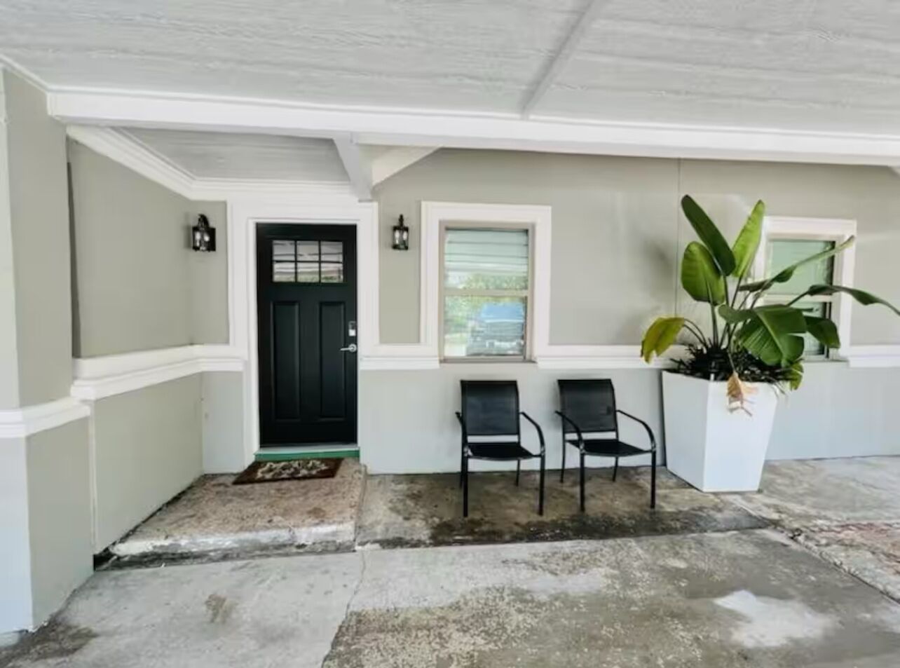 A covered porch with a black door, two chairs, a potted plant, and a small window.