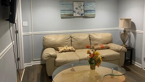 Una sala de estar con un sofá de cuero color beige, una mesa de centro de cristal con un centro de mesa floral, y dos cuadros colgados en la pared que representan paisajes.