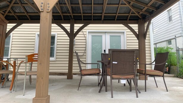 Un patio techado con vigas de madera y cubierta metálica, que incluye mobiliario exterior y una casa al fondo.