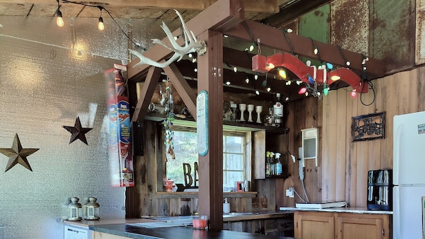 A rustic wooden bar area with stools, a refrigerator, and various decorations including stars and a deer head. - Coastal Escape: Cottage Complex (Brackley Beach)