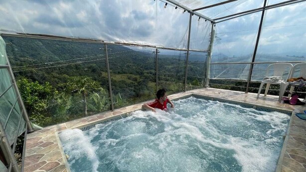 A hot tub with a child in it, surrounded by a glass enclosure with a view of a mountainous landscape. - Finca Recreativa los Altos Giraldos (Arbeláez)