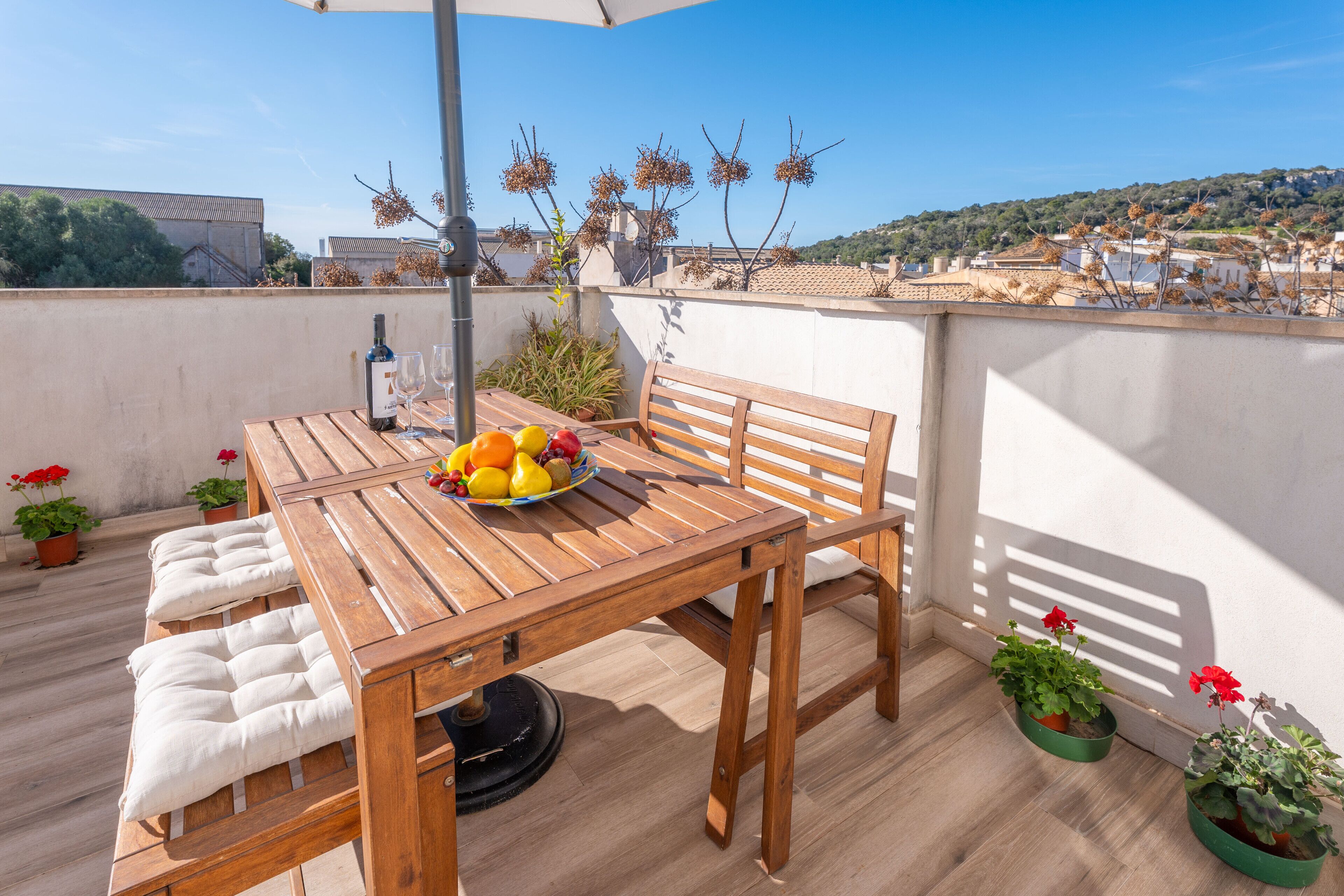 Une table en bois sur laquelle se trouvent un saladier de fruits, une bouteille et des verres, le tout placé sur un balcon agrémenté de plantes en pot et offrant une vue sur des immeubles.