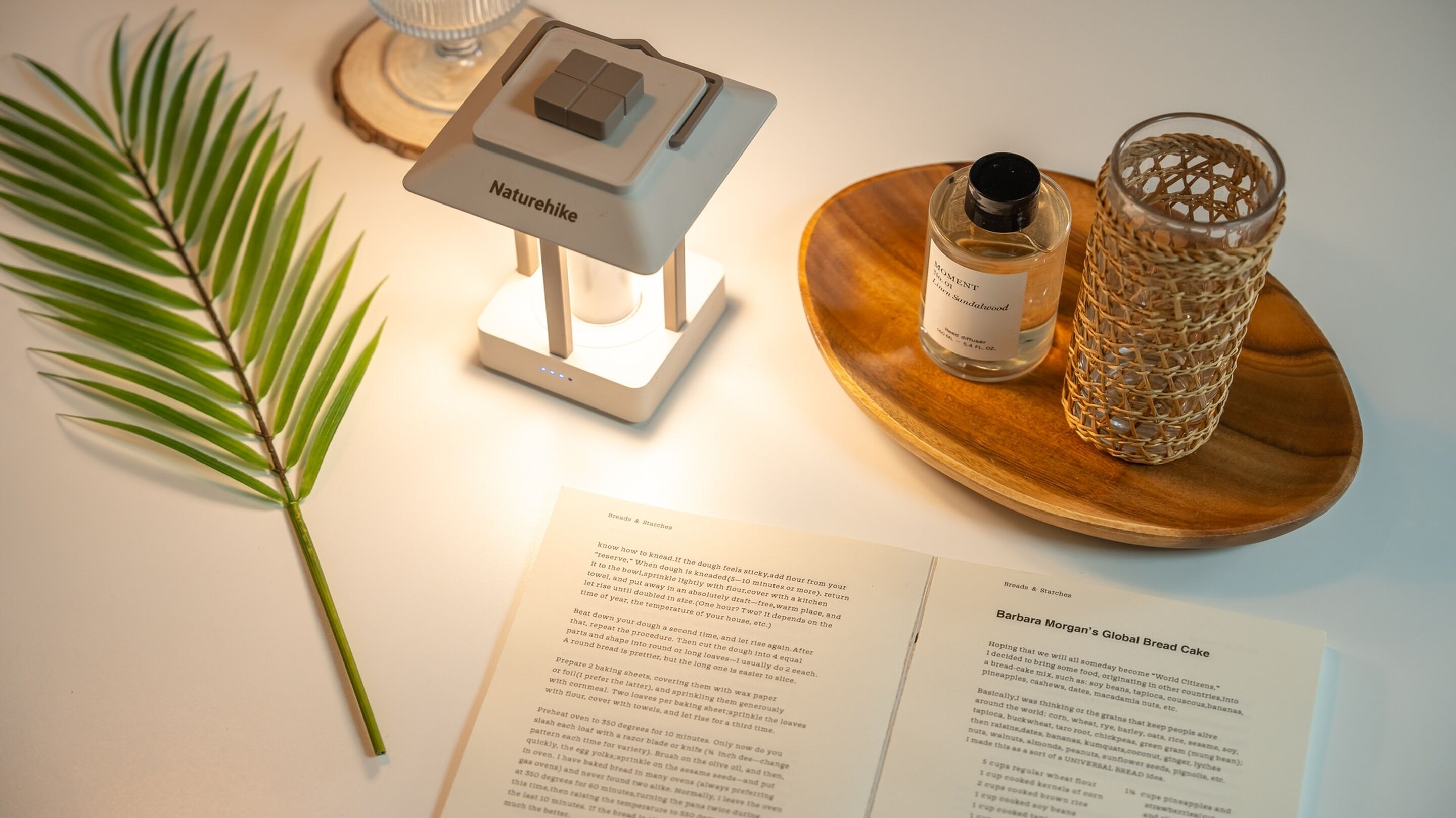 Een keukentablet met een Naturehike apparaat, een fles Moment in Sandalwood, een geweven glazen houder, een palmblad en een kookboek geopend op een pagina met de titel 'Global Bread Cake'.