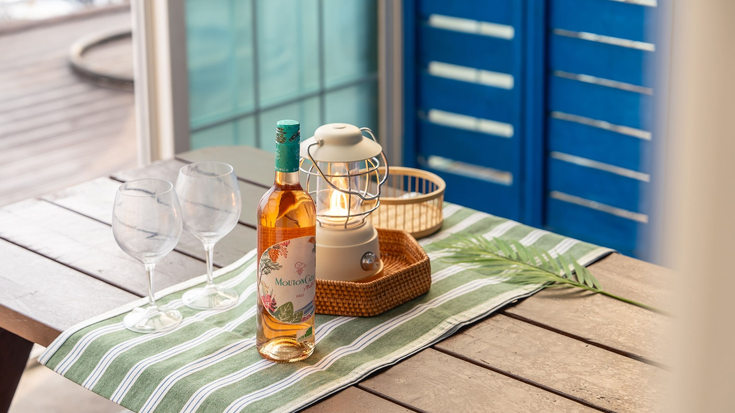 A bottle of Mouton Rothschild wine, two empty wine glasses, a white lantern, and a woven basket on a wooden table with a striped cloth.