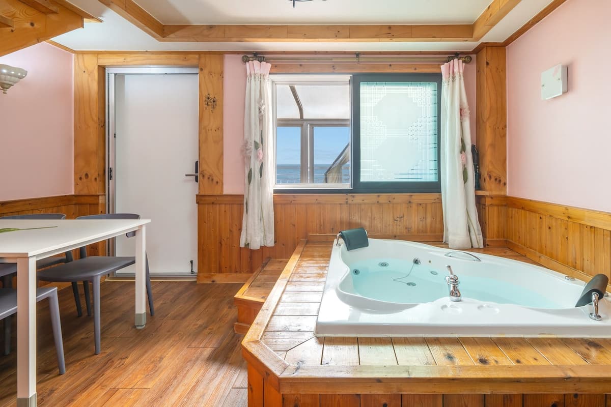 A room with a wooden floor, a white bathtub, a table with chairs, and a window with curtains.