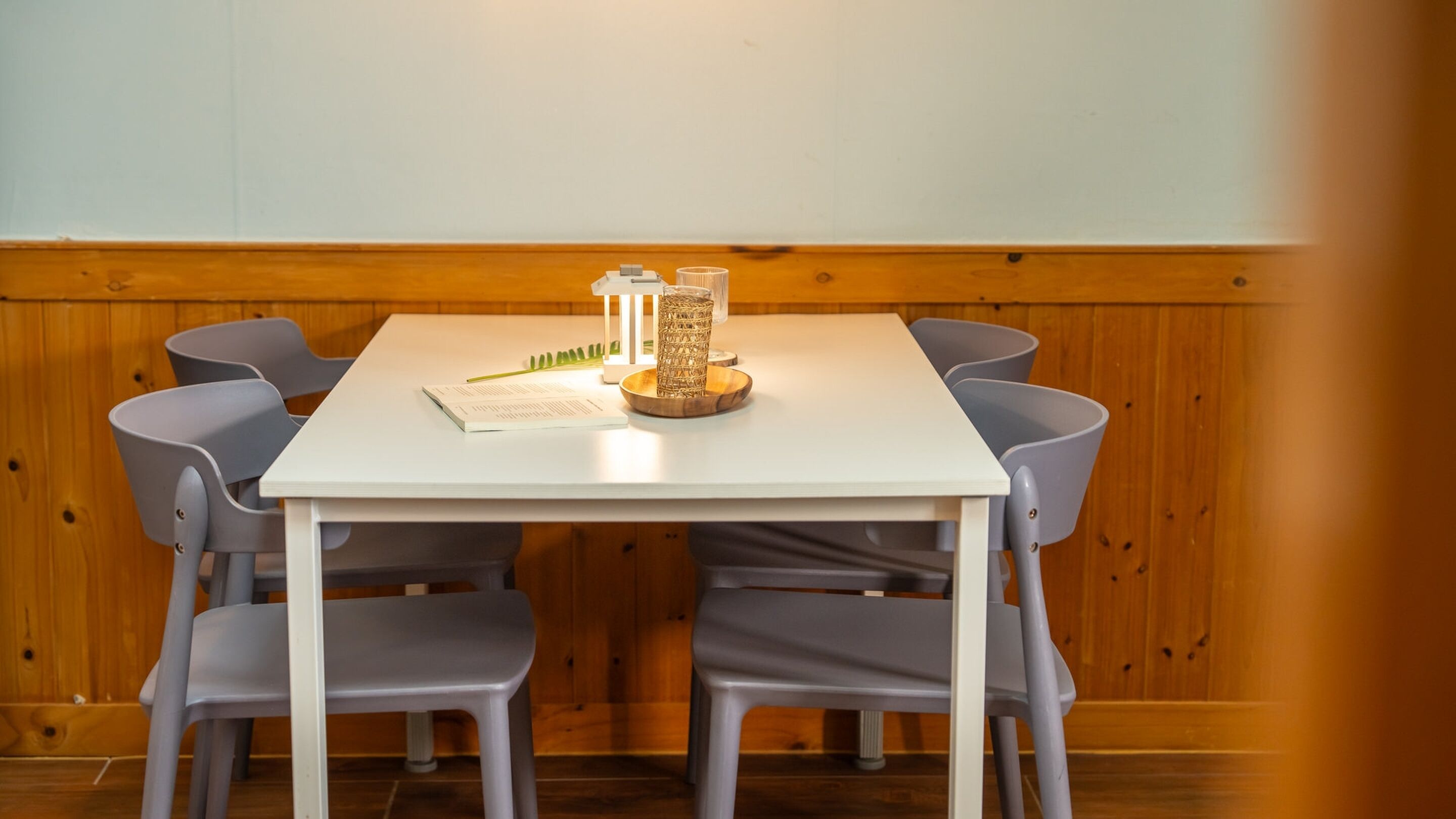 A white table with a candle, a book, and a green plant in a room with wooden paneling and gray chairs.
