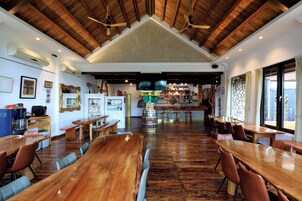 Un comedor espacioso con mesas y sillas de madera, techo alto con vigas de madera y una cocina al fondo.