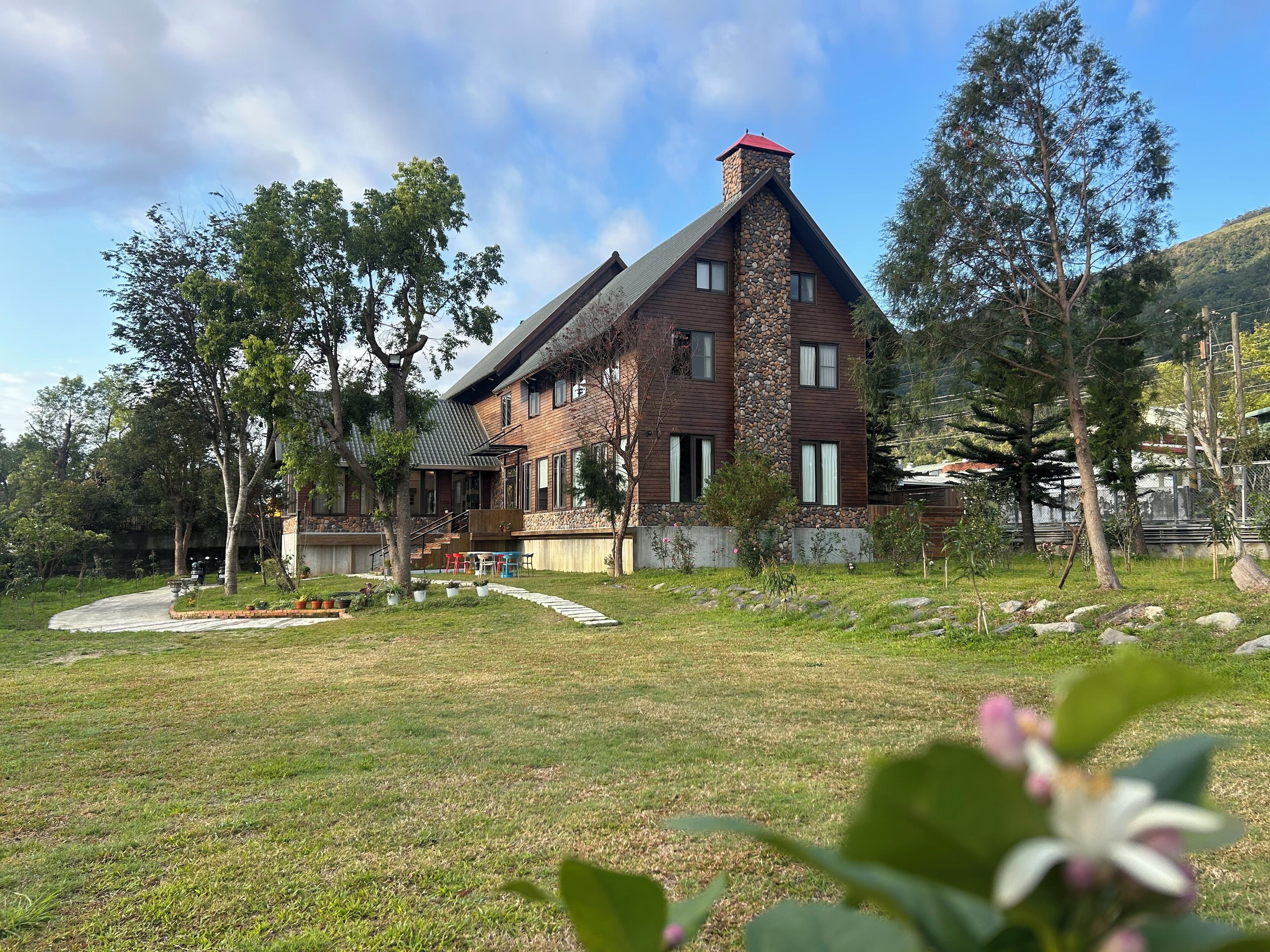 Une maison à deux étages avec une cheminée en pierre, une terrasse en bois et une pelouse bien entretenue.