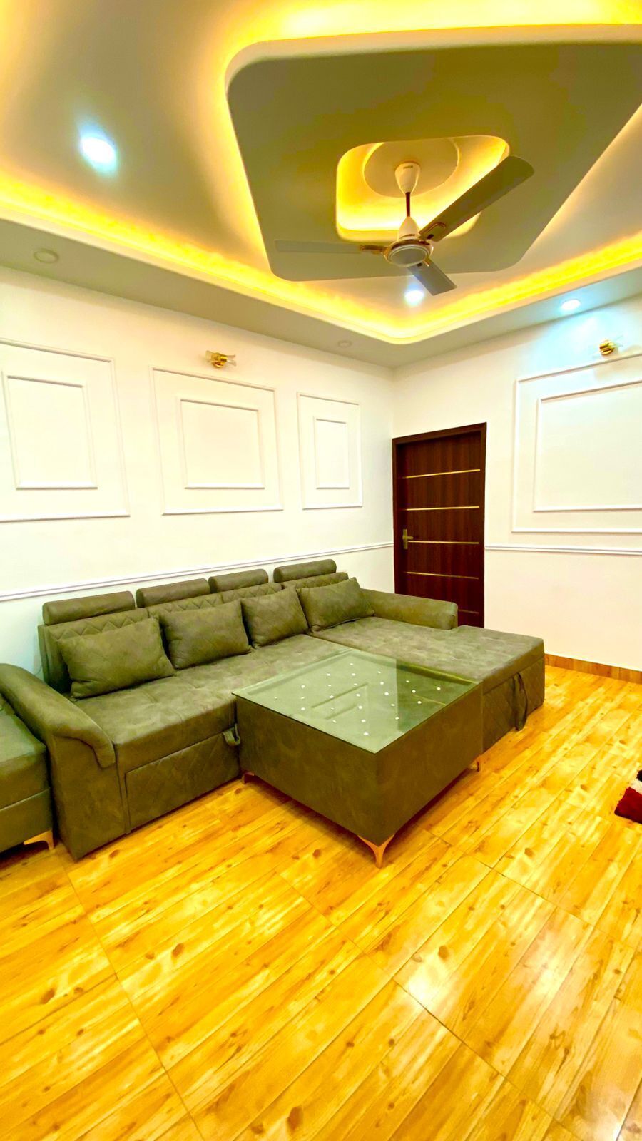 A room with a sectional sofa, wooden flooring, a ceiling fan, recessed lighting, and a dark wooden door.