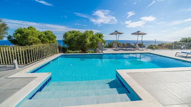 Une piscine d’un bleu éclatant, avec des marches permettant d’y accéder, entourée de chaises longues et de parasols, et offrant une vue sur la mer et les arbres en arrière-plan.
