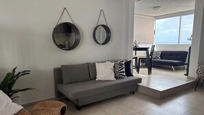 A modern living room with a grey sofa, two round mirrors, a dining area with a table and chairs, and a view through a large window.
