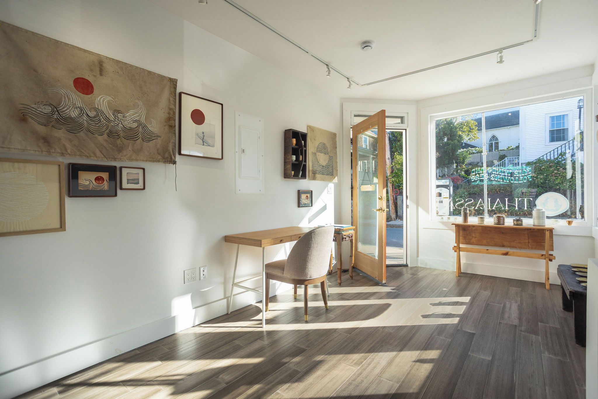 A room with a wooden desk, a chair, and a bench. There are artworks on the walls, a window with a view, and a door leading outside.
