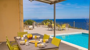 A dining area with a table set for six, surrounded by yellow chairs, overlooking a swimming pool and a scenic view of the sea and mountains. - Villa Dinos with swimming pool (Kefallonia)