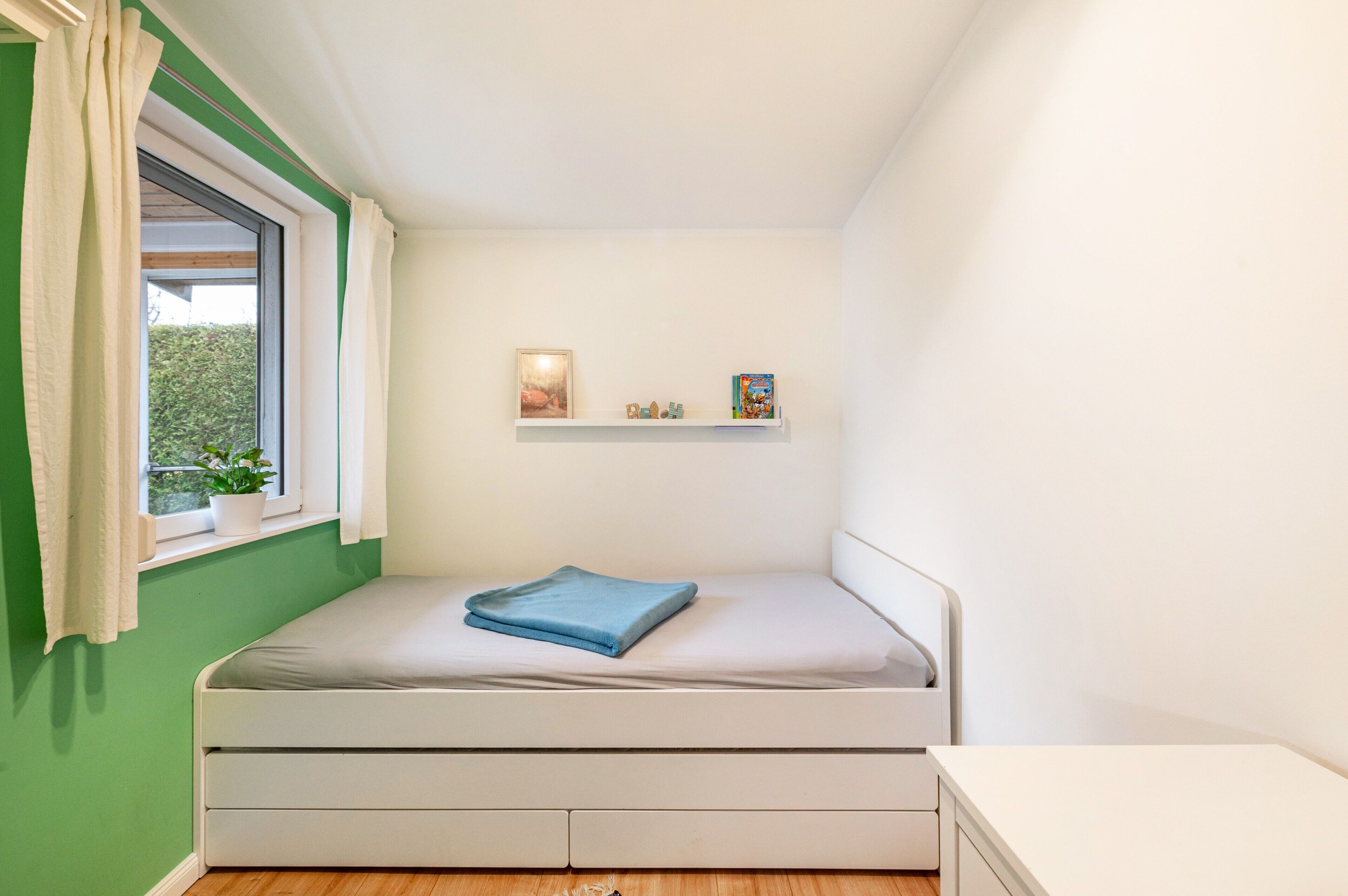 Ein kleines Schlafzimmer mit einem Einzelbett, einem Fenster mit Vorhängen, einem Regal mit Gegenständen und Blick auf Grünfläche.