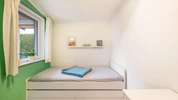 Ein kleines Schlafzimmer mit einem Einzelbett, einem Fenster mit Vorhängen, einem Regal mit Gegenständen und Blick auf Grünfläche.