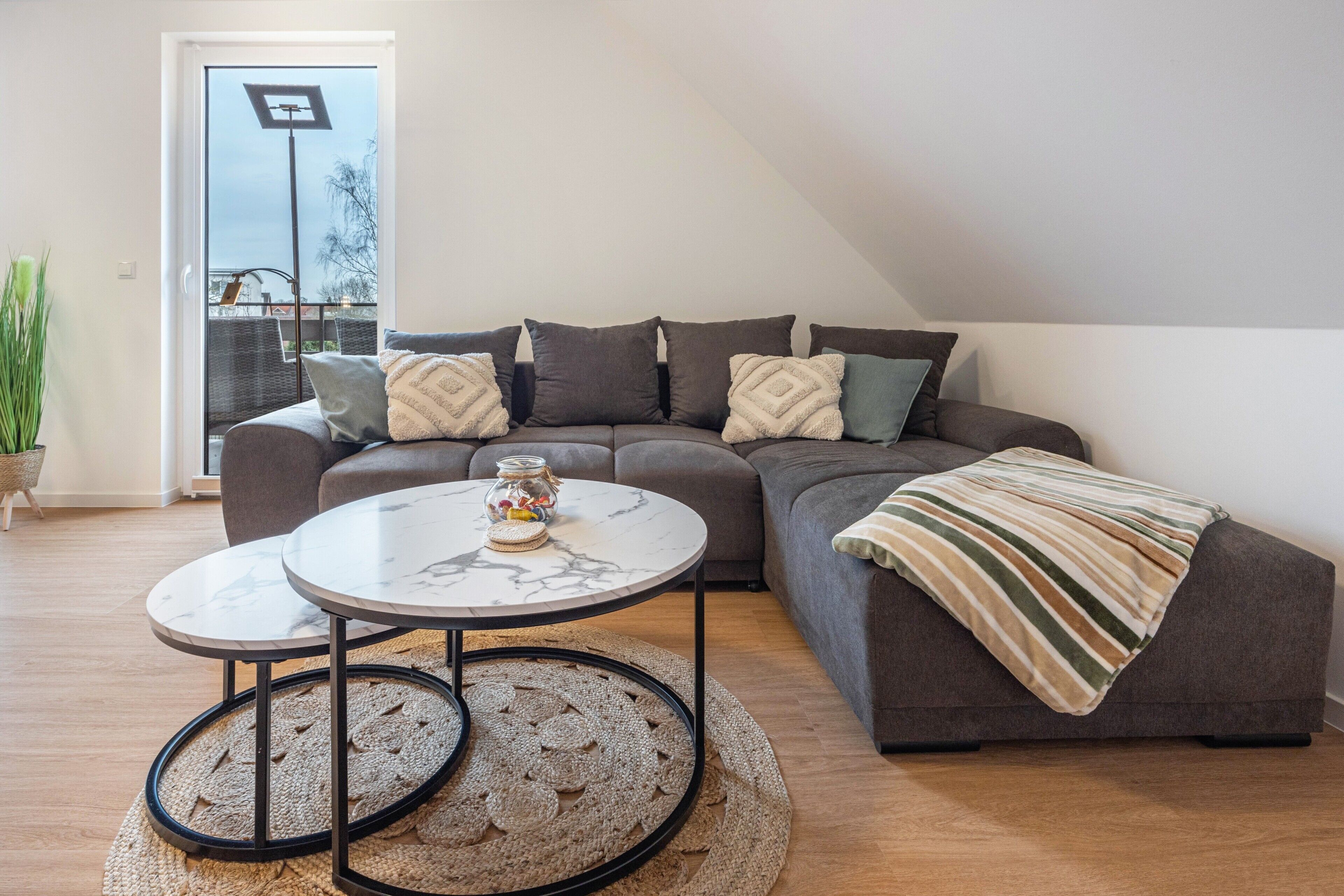 Un salon moderne avec un canapé d’angle, deux tables rondes, un tapis tissé et une plante en pot.