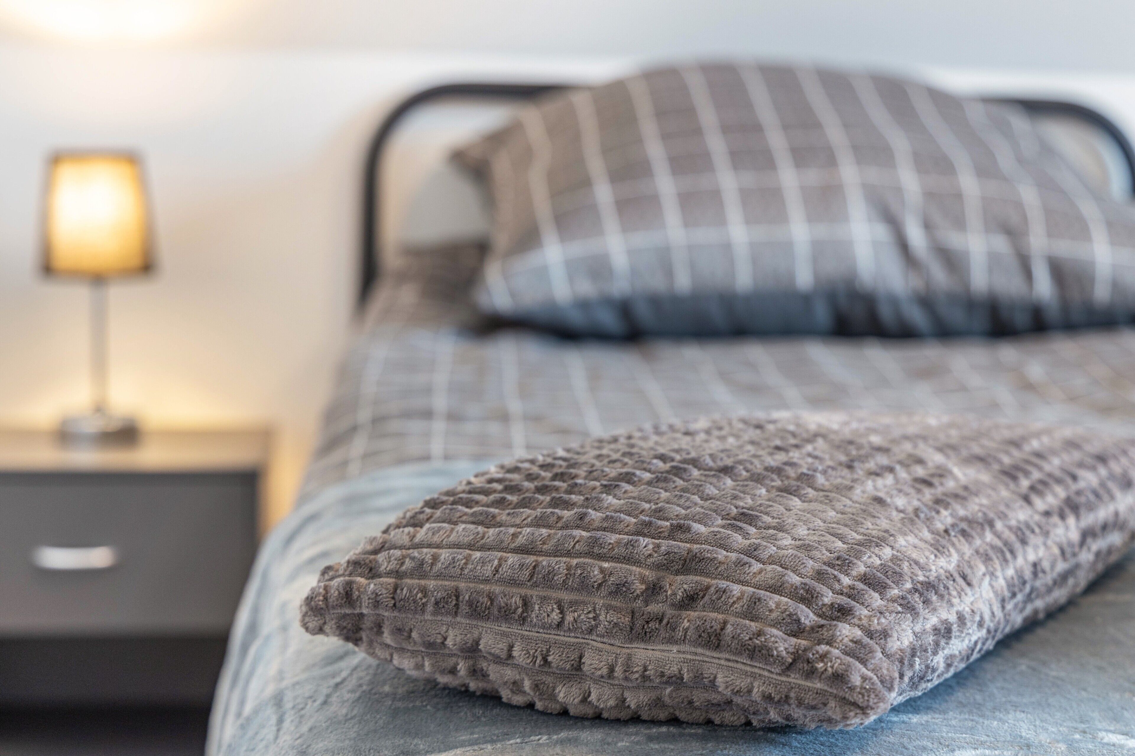 Una cama con un edredón con textura y una almohada a cuadros, una lámpara de noche y una mesita de noche.