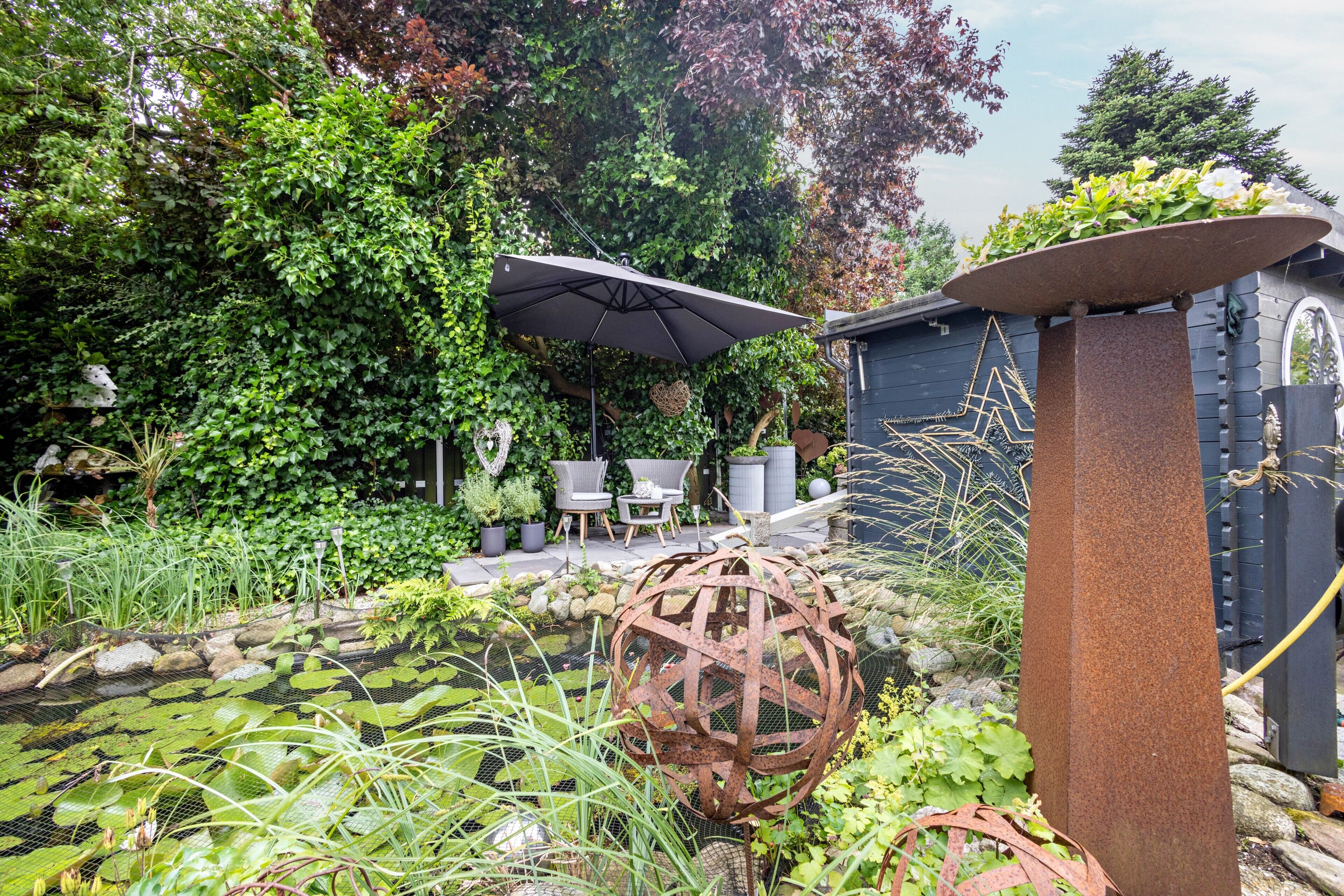 A garden with a pond, a metal sculpture, and a seating area with chairs and an umbrella.