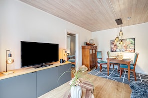 Ein modernes Wohnzimmer mit Flachbildfernseher, Holzboden, Esstisch mit Stühlen und einer Vase mit Blumen.