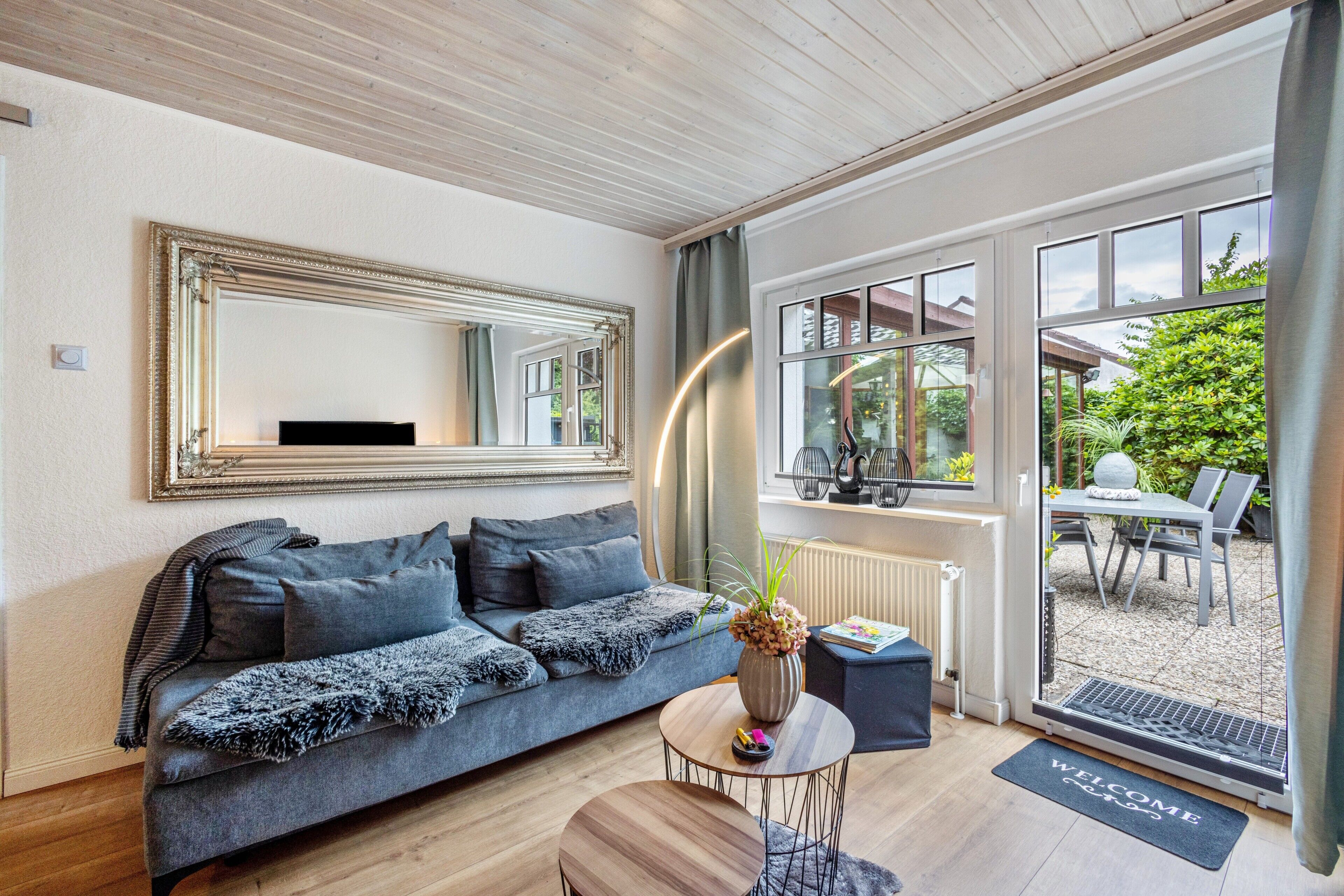 A modern living room with a sofa, a round coffee table, a large mirror, and a view of the garden through glass doors.