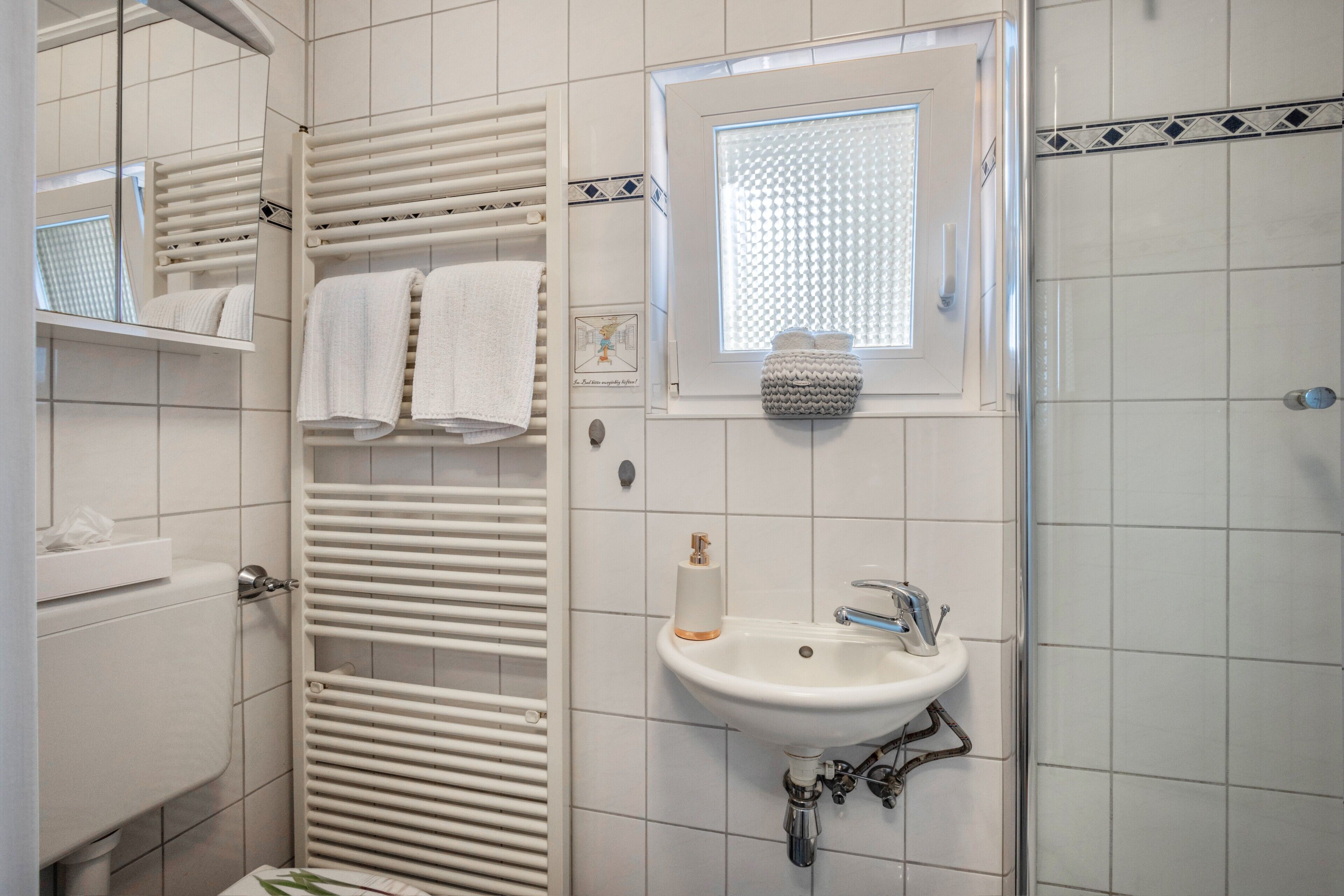 Un baño con piso de baldosas blancas, un lavamanos blanco con dispensador de jabón, un perchero con dos toallas, una ventana con persiana texturizada y una zona de ducha.