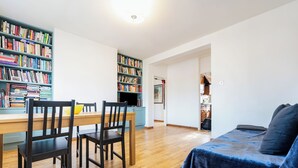 A room with a wooden dining table, chairs, a bookshelf, a bed with a blue blanket, and a kitchen area in the background. - 2 bedroom flat in Hackney, London. Next to several stations and restaurants (London)