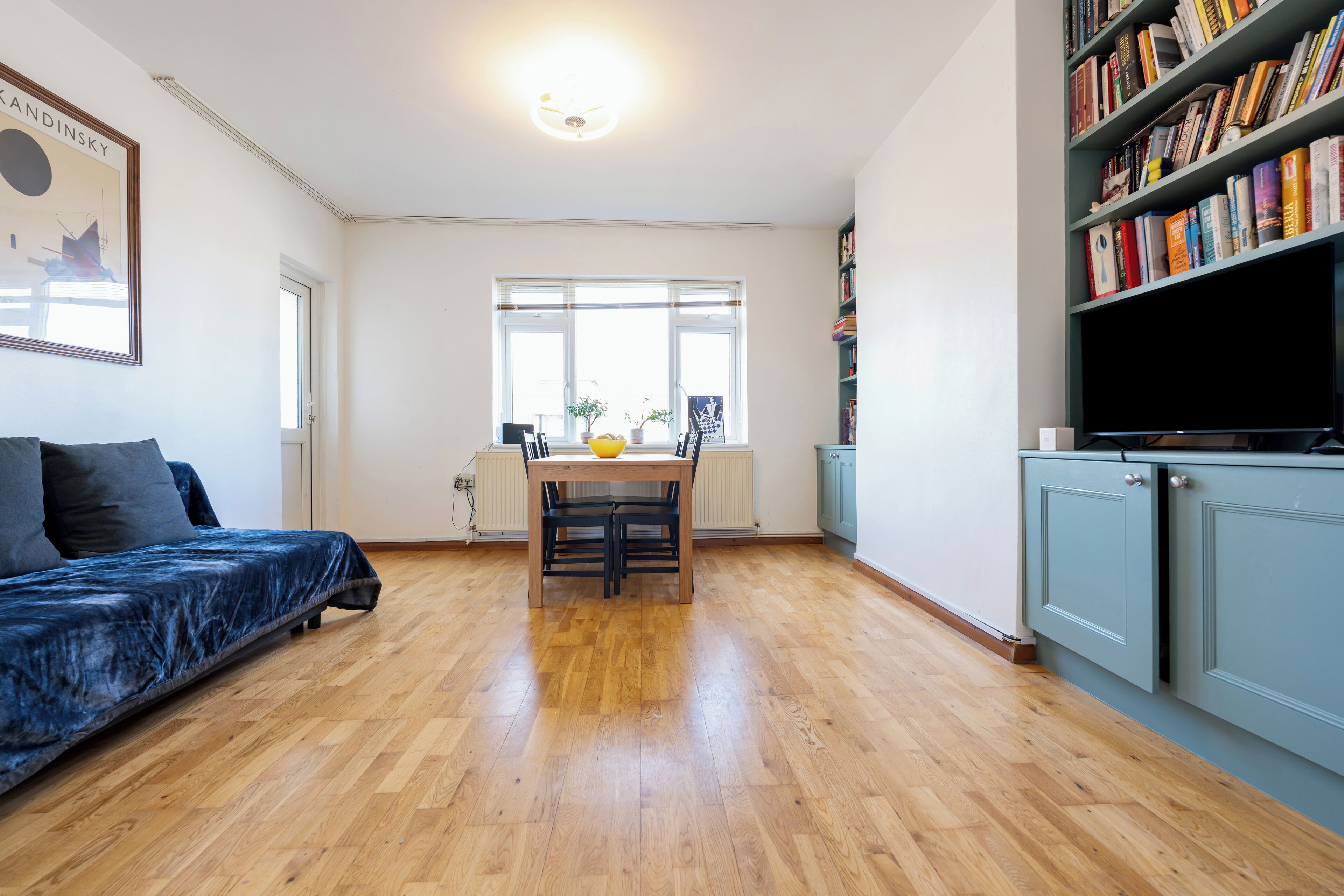 A spacious living room with a wooden floor, a blue sofa, a dining table with chairs, a bookshelf, and a television.