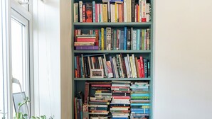 A room with a wooden table, a bookshelf filled with books, and a window with blinds. - 2 bedroom flat in Hackney, London. Next to several stations and restaurants (London)