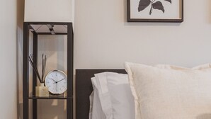 Een slaapkamer met een bed, nachtkastje, klok, plant in een lijst en een afvalbak.