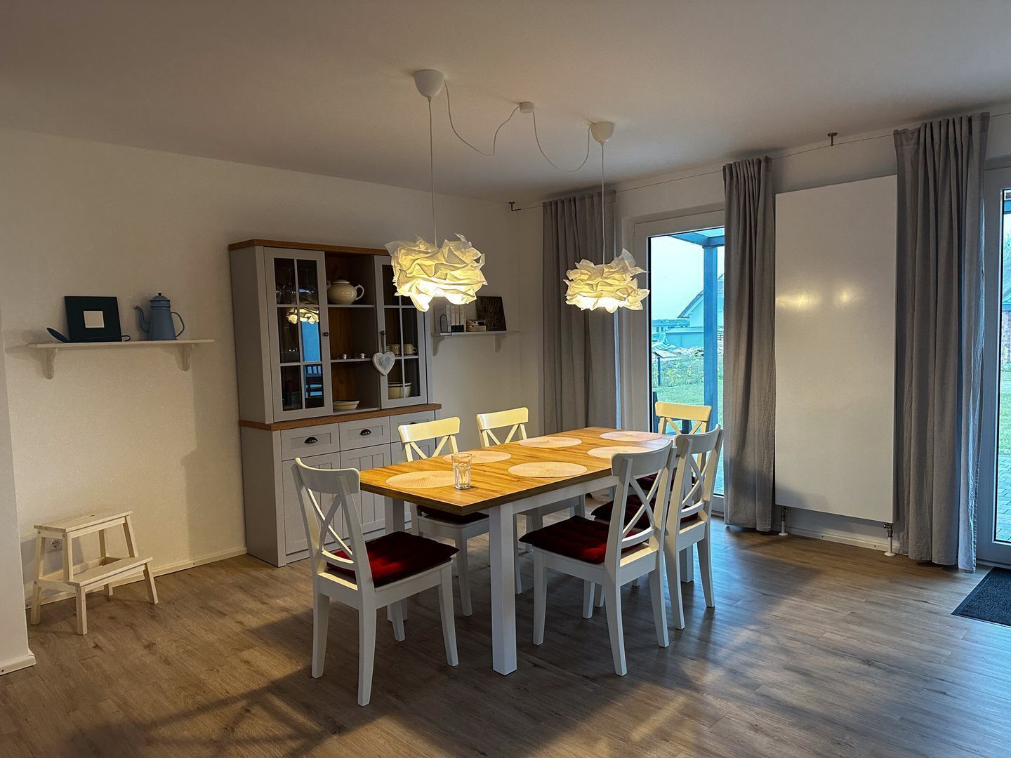 A dining area with a wooden table, white chairs with red cushions, a cabinet with glass doors, and a view through large windows.