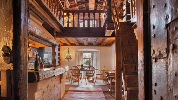 Un interior rústico con vigas de madera, un comedor con sillas, una ara ña y vistas a través de un ventanal.