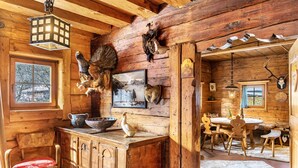 Interior rústico de una cabaña con paredes de madera, una zona de comedor con mesa y sillas, una chimenea y varias cabezas de animales decorativas.