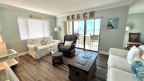 Un salón con vistas a la playa, un sofá blanco, una silla de mimbre y una mesa de centro de madera.