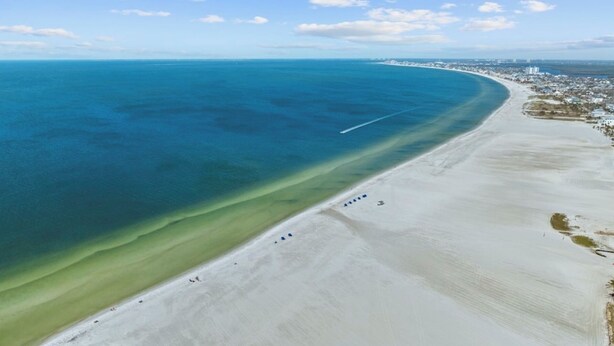Vista aérea de una playa con un océano azul cristalino, una orilla arenosa y un paisaje urbano distante.