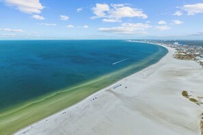 An aerial view of a beach with a clear blue ocean, a sandy shore, and a distant cityscape.