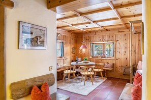 Interior de una cabaña de madera con una mesa de comedor, sillas, un banco, una lámpara colgante y una ventana con cortinas.