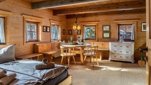 Interior de una cabaña de madera con una cama, asientos en un banco, una mesa de comedor con sillas, un aparador y cuadros en las paredes.