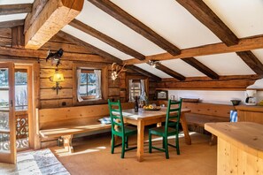 A rustic wooden cabin with a dining area featuring a wooden table and green chairs, a bench, and a kitchen area with a wooden counter.