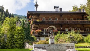 Ein hölzernes Gebäude mit einem Balkon, umgeben von Grünpflanzen und einem Steinmonument.