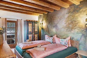 A room with two beds, a wooden ceiling, a mural, and a window with curtains.