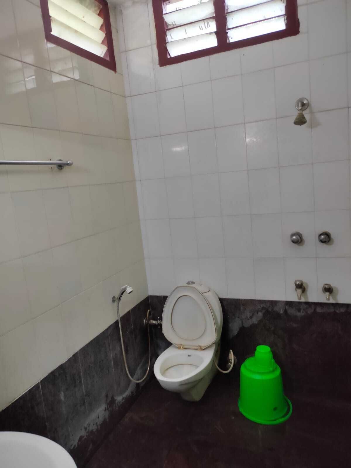 Une salle de bain équipée de toilettes, d’une barre d’appui, d’une poubelle verte et d’une fenêtre avec des stores.