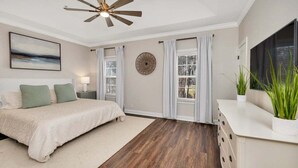Une chambre à coucher avec un lit, un ventilateur de plafond, une grande fenêtre avec des rideaux, une horloge murale et une plante.