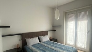 Une chambre à coucher comprenant un lit, des tables de chevet, un luminaire suspendu et une fenêtre avec des stores.